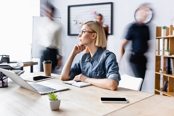 Rubia gerente mirando hacia otro lado mientras está sentado en el escritorio con gadgets y café para ir en la oficina de espacio abierto, desenfoque de movimiento - foto de stock