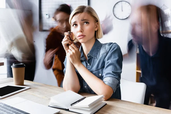 Traurige Geschäftsfrau mit Brille in der Nähe von Notizbuch im Großraumbüro sitzend, Bewegungsunschärfe — Stockfoto