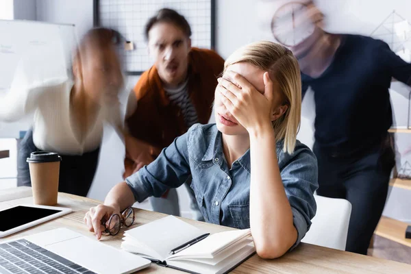 Gerente cansado obscurecendo rosto com a mão enquanto sentado perto notebook no escritório espaço aberto, borrão de movimento — Fotografia de Stock