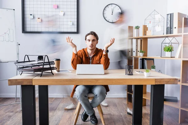 Confused businessman showing shrug gesture while sitting at desk in open space office, motion blur — Stock Photo