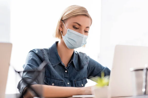 Rubia manager en médico máscara trabajo en portátil en borrosa primer plano - foto de stock
