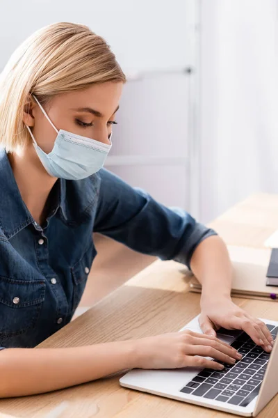 Jeune gestionnaire en masque médical tapant sur ordinateur portable dans le bureau — Photo de stock