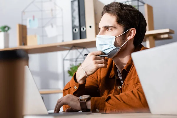 Pensativo hombre de negocios en máscara médica mirando mientras sostiene la pluma en primer plano borrosa - foto de stock