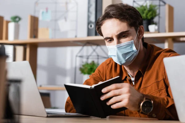 Jovem empresário em máscara médica olhando no notebook no local de trabalho em fundo embaçado — Fotografia de Stock
