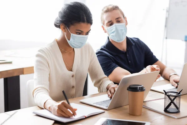 Mujer de negocios afroamericana escribiendo en el ordenador portátil una escritura en el cuaderno cerca de colega en máscara médica sobre fondo borroso - foto de stock