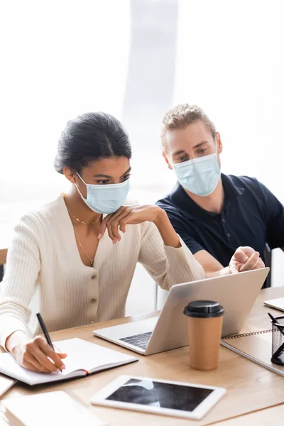 African american businesswoman in medical mask near manager pointing with finger at laptop — Stock Photo