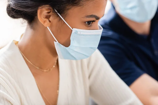 Mujer de negocios afroamericana en máscara médica cerca de colega en fondo borroso - foto de stock