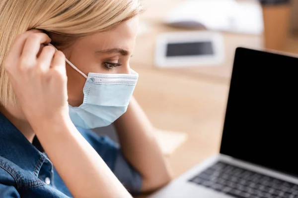 Joven gerente poner máscara médica en la cara cerca de la computadora portátil sobre fondo borroso - foto de stock