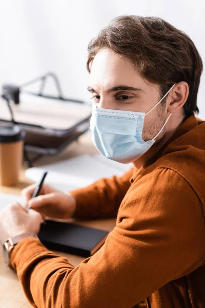 Молодий менеджер в медичній масці сидить на робочому місці з ручкою і блокнотом на розмитому фоні — стокове фото