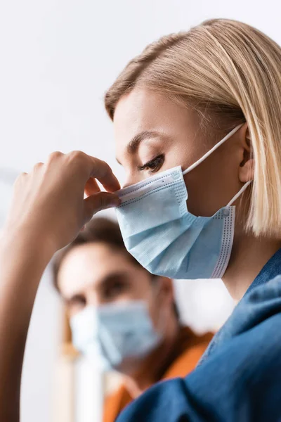 Blonde businesswoman fixing medical mask near colleague on blurred background — Stock Photo