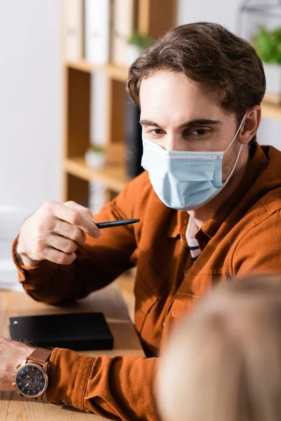 Менеджер в медичній масці вказує з ручкою, сидячи на робочому місці — стокове фото