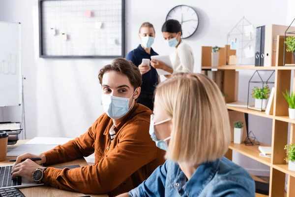 Joven gerente en máscara médica mirando a colega mientras que los empresarios hablando sobre fondo borroso - foto de stock