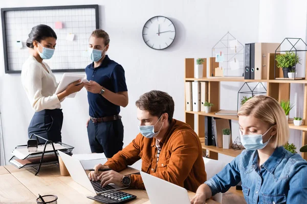 Empresarios en máscaras médicas que trabajan en computadoras portátiles cerca de colegas multiétnicos hablando mientras sostiene gadgets - foto de stock