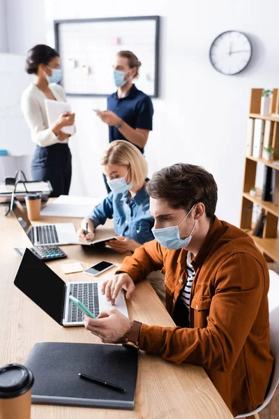Giovani imprenditori multiculturali in maschere mediche che lavorano in ufficio all'aperto durante la quarantena su sfondo sfocato — Foto stock