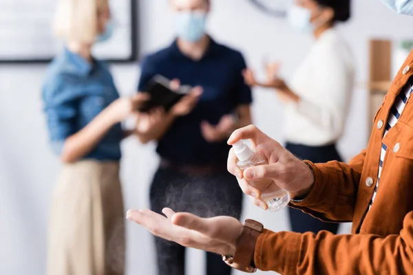 Vista ritagliata di uomo d'affari spruzzando disinfettante sulle mani mentre i colleghi parlano su sfondo sfocato — Foto stock