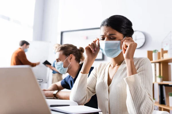 Disgustado afroamericano gerente poner en máscara médica cerca de colegas multiculturales que trabajan en fondo borroso - foto de stock