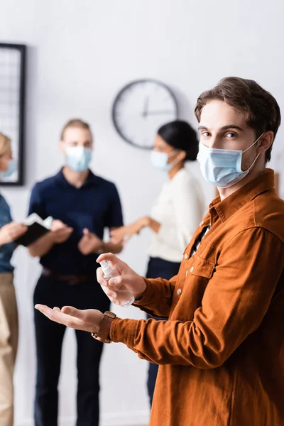 Jovem empresário em máscara médica segurando desinfetante enquanto olha para a câmera perto de colegas em fundo borrado — Fotografia de Stock