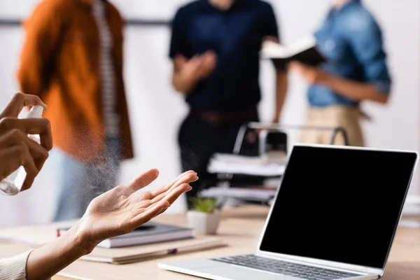 Vista ritagliata di donna d'affari afro-americana spruzzando antisettico su mani vicino laptop e colleghi su sfondo sfocato — Foto stock