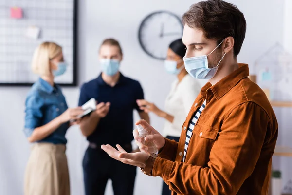 Gerente em máscara médica pulverização desinfetante em mãos perto de colegas em fundo turvo — Fotografia de Stock