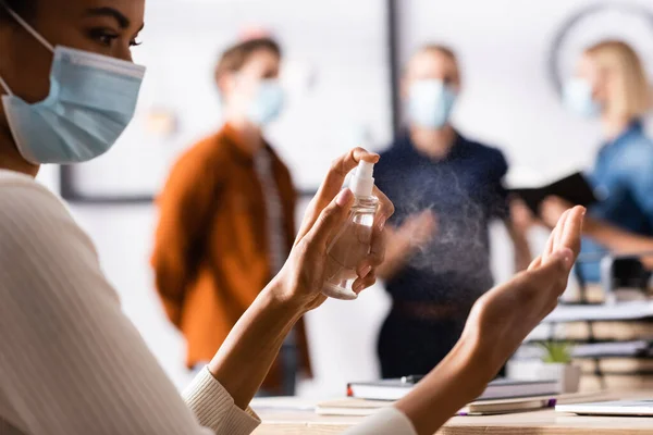 Partial view of african american manager disinfecting hands at workplace, and colleagues  standing on blurred background — Stock Photo