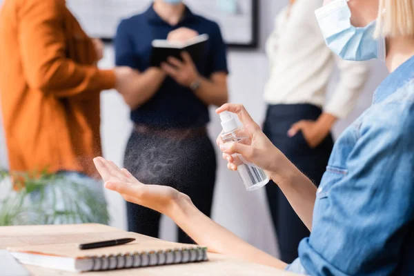 Abgeschnittene Ansicht einer Geschäftsfrau in medizinischer Maske, die antiseptische Mittel auf Hände in der Nähe von Kollegen auf verschwommenem Hintergrund sprüht — Stockfoto