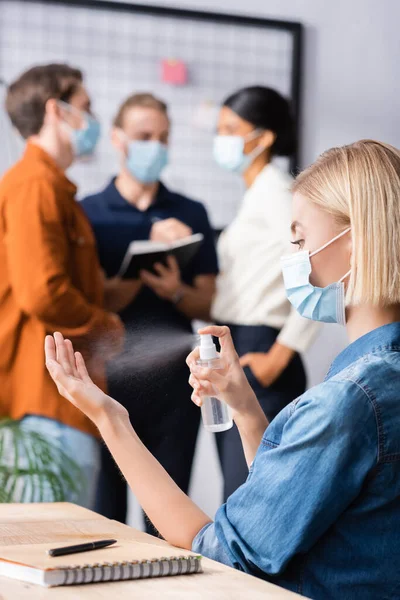 Rubia mujer de negocios en máscara médica rociando desinfectante cerca de colegas sobre fondo borroso - foto de stock