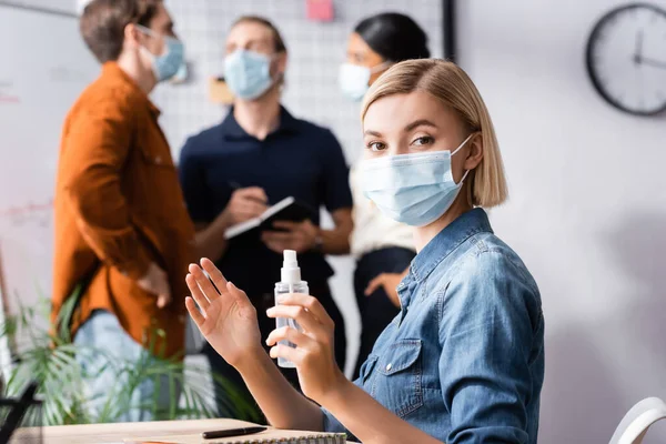 Manager bionda in maschera medica che tiene il disinfettante mentre guarda la fotocamera vicino ai colleghi su sfondo sfocato — Foto stock