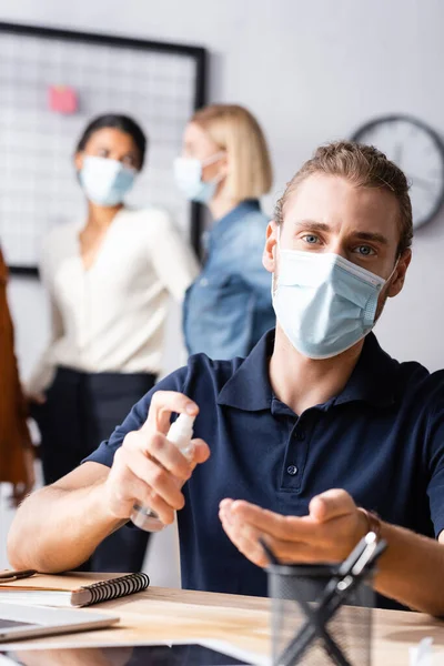 Gerente em máscara médica pulverizando desinfetante em mãos enquanto olha para a câmera perto de colegas falando em fundo borrado — Fotografia de Stock
