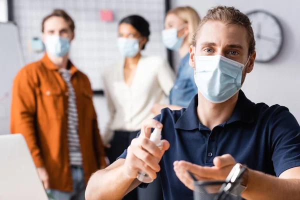 Joven hombre de negocios en máscara médica rociando desinfectante en las manos cerca de colegas hablando sobre fondo borroso - foto de stock