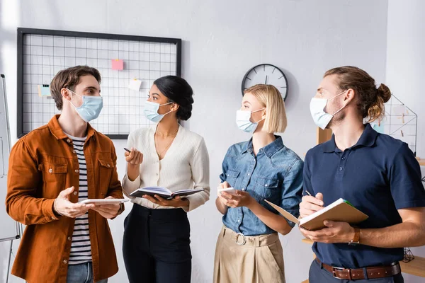 Jóvenes empresarios multiculturales en máscaras médicas discutiendo proyecto de negocio - foto de stock