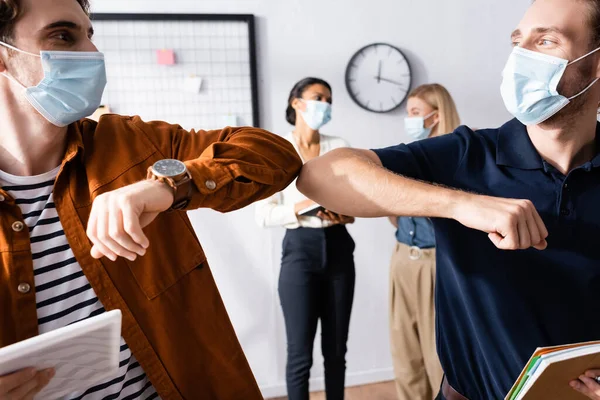 Junge Manager in medizinischer Maske machen Ellenbogenschlag-Geste in der Nähe von Geschäftsfrauen auf verschwommenem Hintergrund — Stockfoto