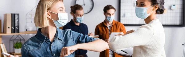 Jóvenes mujeres de negocios en máscaras médicas haciendo golpe de codo cerca de colegas sobre fondo borroso, pancarta - foto de stock