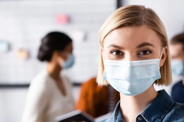 Blonde Geschäftsfrau in medizinischer Maske blickt in die Kamera, während multiethnische Kollegen über verschwommenen Hintergrund sprechen — Stockfoto