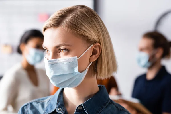 Jovem, gerente loira em máscara médica olhando para longe perto turva colegas multiétnicos em segundo plano — Fotografia de Stock