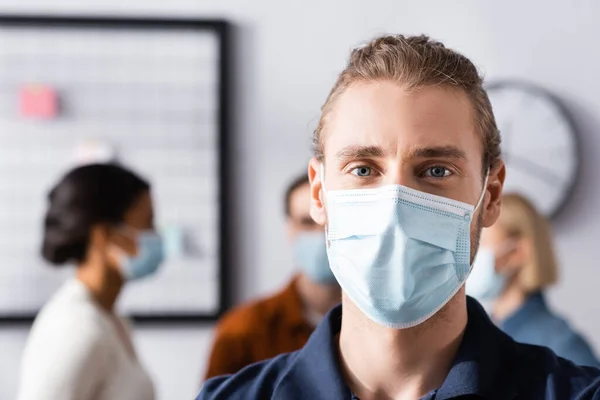 Young office manager in medical mask looking at camera near multicultural colleagues on blurred background — Stock Photo