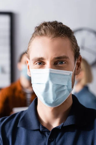 Молодий менеджер в медичній масці дивиться на камеру на розмитому фоні — стокове фото