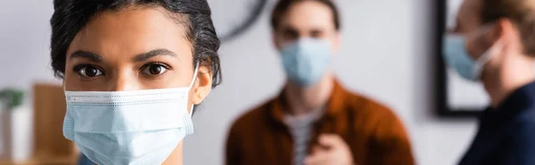 African american businesswoman in protective mask looking at camera near blurred managers on background, banner — Stock Photo