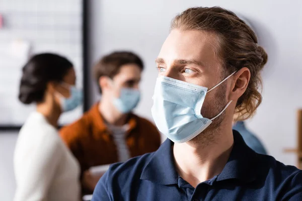Hombre de negocios con máscara médica mirando hacia otro lado mientras colegas multiculturales hablando sobre fondo borroso - foto de stock