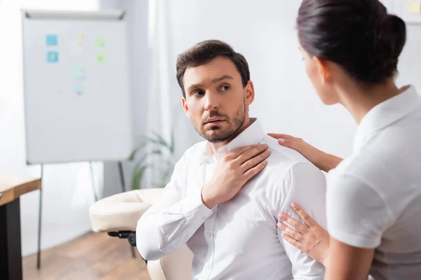 Uomo d'affari con dolore alla spalla guardando massaggiatrice in ufficio su sfondo sfocato — Foto stock