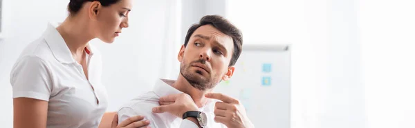 Masajista mirando al hombre de negocios señalando con el dedo al hombro doloroso en la oficina sobre fondo borroso, pancarta - foto de stock