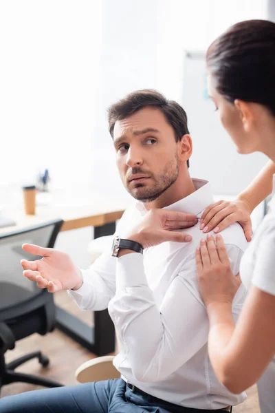 Masseurin massiert schmerzende Schulter eines Geschäftsmannes, der im Büro auf verschwommenem Hintergrund gestikuliert — Stockfoto