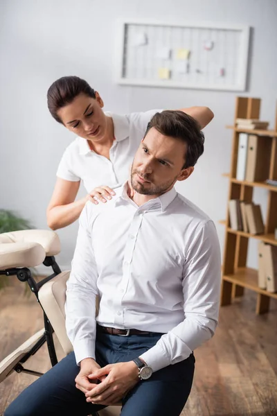 Masajista masajeando hombro de hombre de negocios sentado en silla de masaje en la oficina sobre fondo borroso - foto de stock