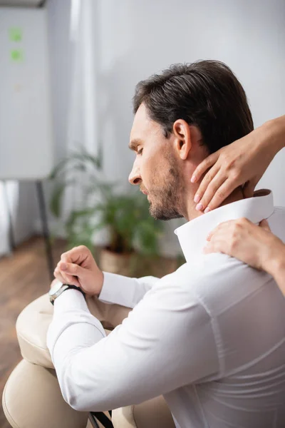 Massage-Therapeut massiert schmerzhaften Nacken des Kunden sitzt auf Massagesessel im Büro auf verschwommenem Hintergrund — Stockfoto