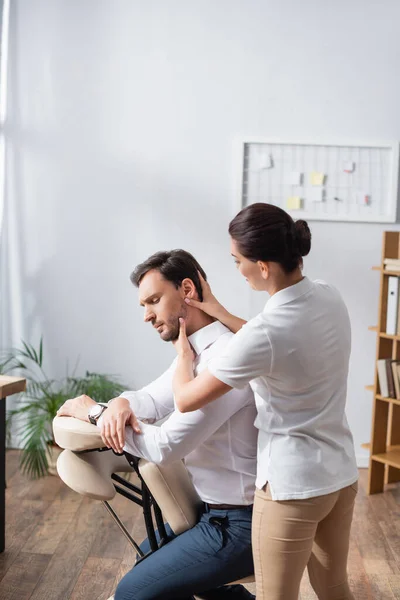 Massaggiatrice massaggiando testa di uomo d'affari seduto su sedia da massaggio in ufficio su sfondo sfocato — Foto stock