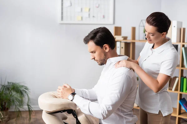 Lächelnde Masseurin massiert Schultern eines ernsthaften Geschäftsmannes, der auf Massagestuhl im Büro auf verschwommenem Hintergrund sitzt — Stockfoto