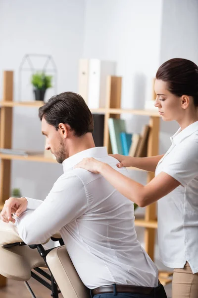 Massagetherapeutin massiert Schultern eines Geschäftsmannes, der auf Massagesessel im Büro auf verschwommenem Hintergrund sitzt — Stockfoto