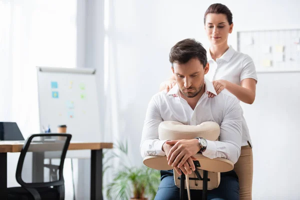 Smiling masseuse massaging shoulders of businessman sitting on massage chair in office on blurred background — Stock Photo