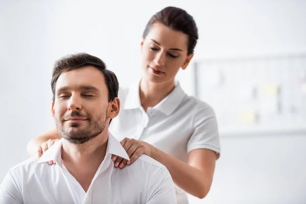 Masajista masajeando hombros de hombre de negocios sonriente con los ojos cerrados sobre fondo borroso - foto de stock