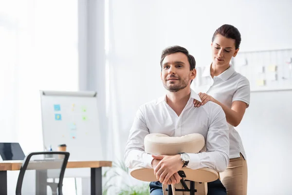 Masajista masajeando hombro de hombre de negocios sonriente sentado en silla de masaje en la oficina sobre fondo borroso - foto de stock