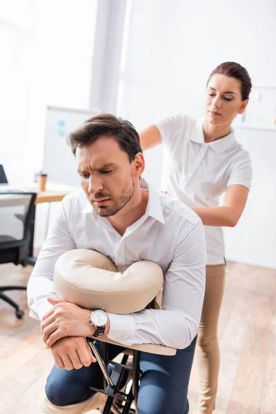 Klient mit Rückenschmerzen sitzt auf Massagesessel, während Masseurin sitzende Massage im Büro auf verschwommenem Hintergrund macht — Stockfoto